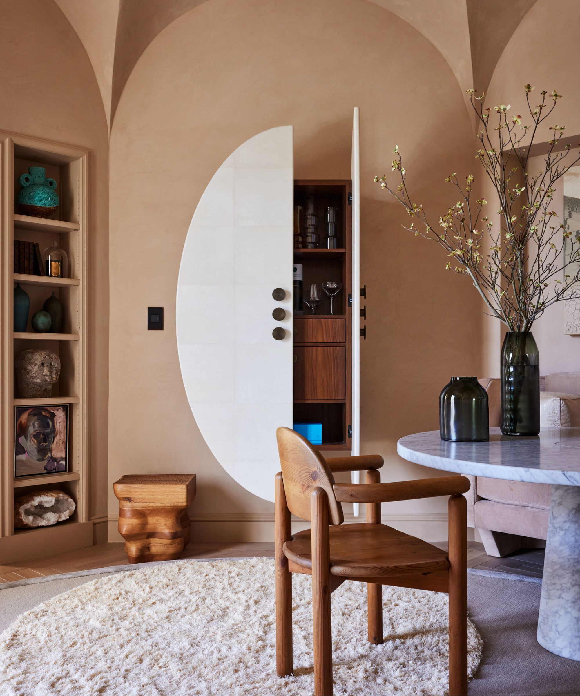 A dining room area with beige walls painted in Adobe Dust by Benjamin Moore. There are doors leading to a cupboard which has a pale off white circle motif painted on them, and a dining area featuring a round marble table, a wooden chair and a round sham rug.