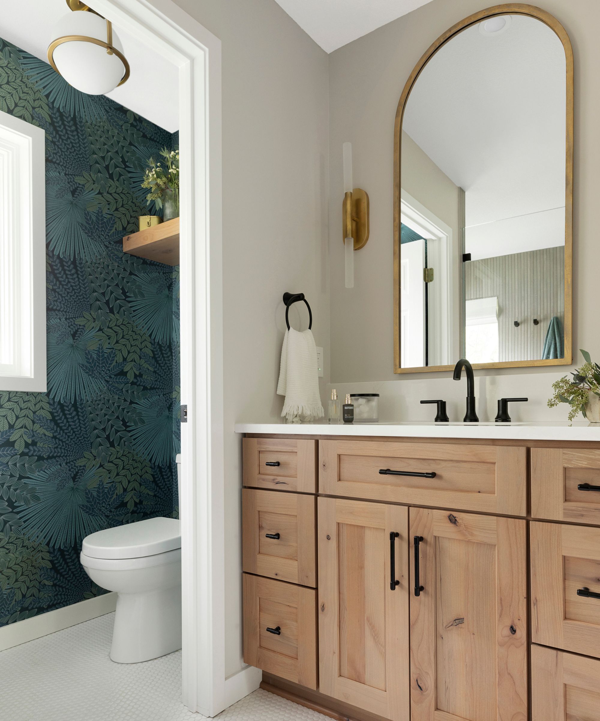 A modern bathroom with a wooden basin vanity unit and an over basin mirror, the walls are painted in Canvas Tan by Sherwin Williams and the W/C has dark green wallpaper with a fern and foliage pattern.