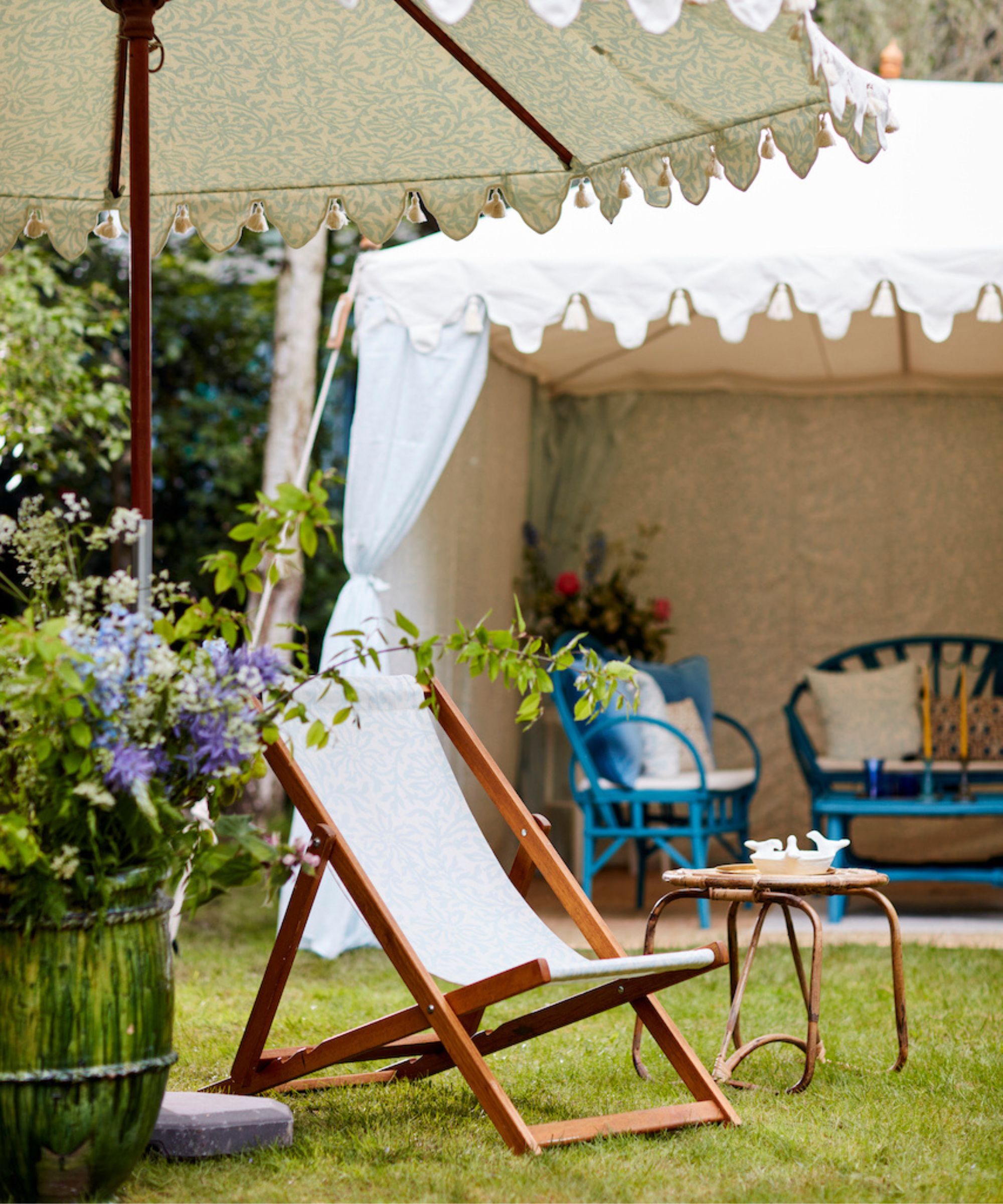Deckchair and barrel of purple flowers under a parasol with a white tent in the background with blue garden furniture inside