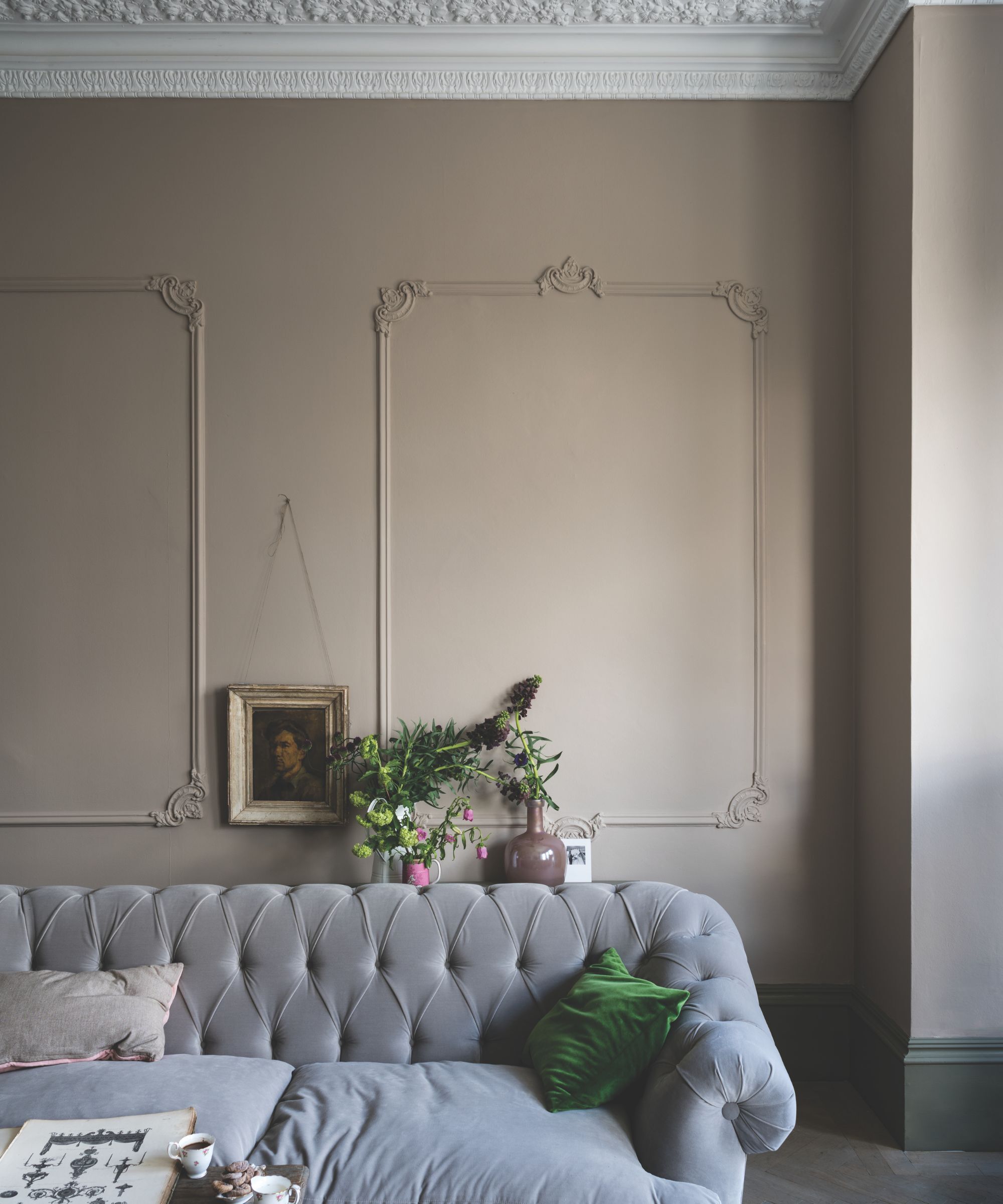 A sitting room with a gray velvet sofa and walls in a Jitney by Farrow & Ball. There is an ornate white ceiling and intricate architectural mouldings on the wall.