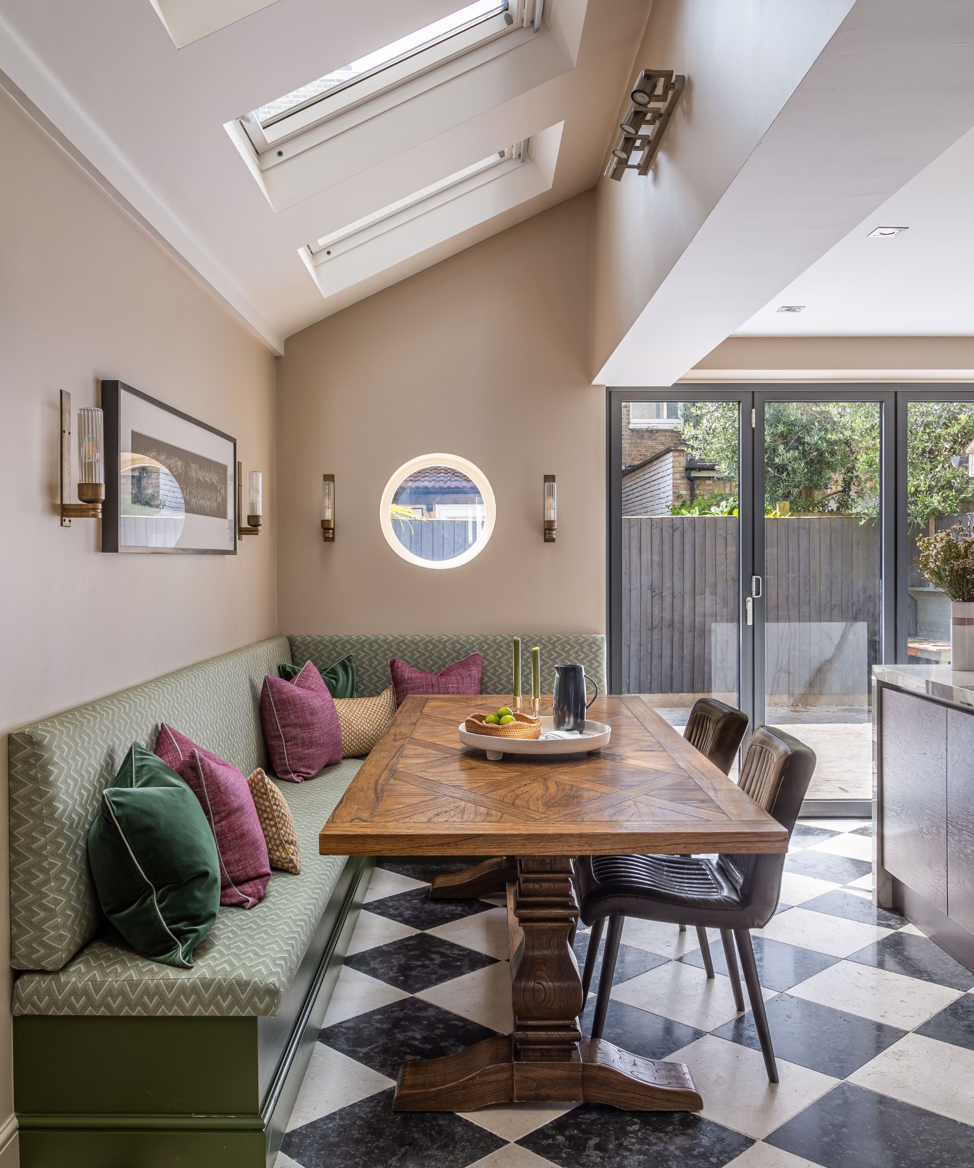 A modern kitchen extension with walls painted in Oxford Stone by Farrow & Ball, a green bench seat and a kitchen dining table, the floor is black and white tiles and there are bi-fold doors to a courtyard garden.