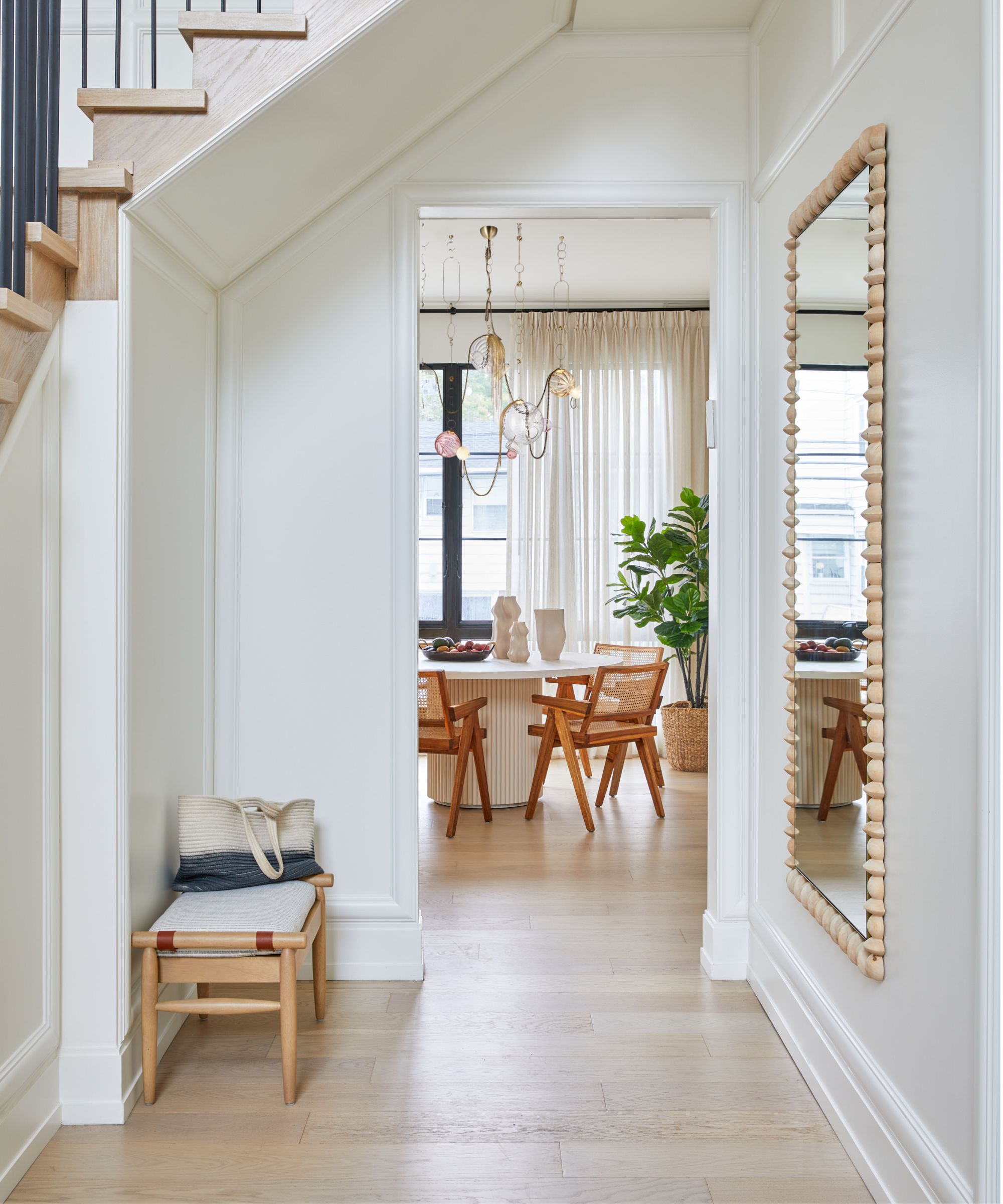 Modern beige entryway leading to a dining room, painted in White Dove by Benjamin Moore. There is a staircase leading upstairs, a mirror on the hallway wall and a small bench with a bag on it in the entryway.
