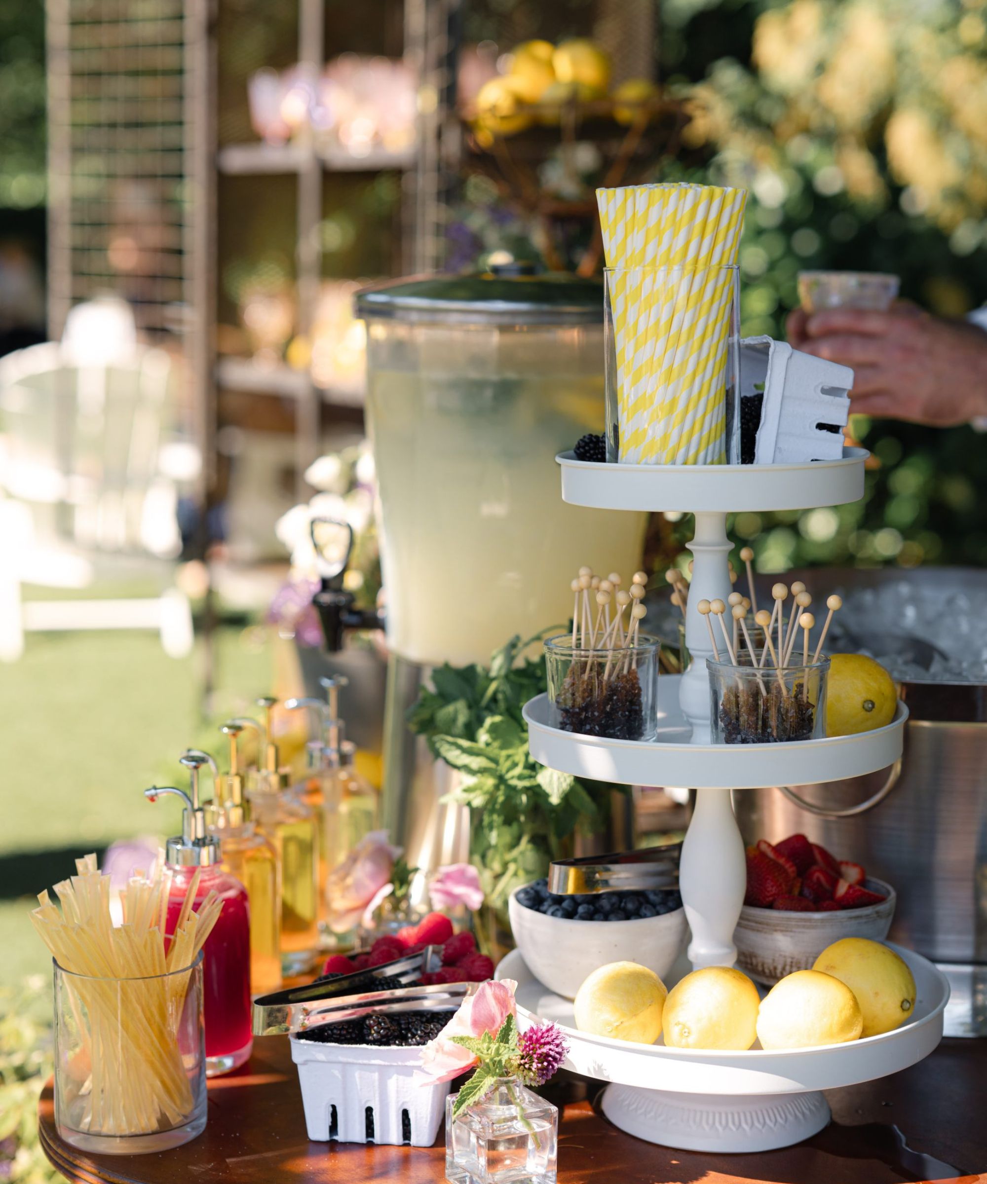 Drinks table set up outdoors with lemonade dispenser and syrups, and tiered trays of lemons, snacks and yellow and white straws