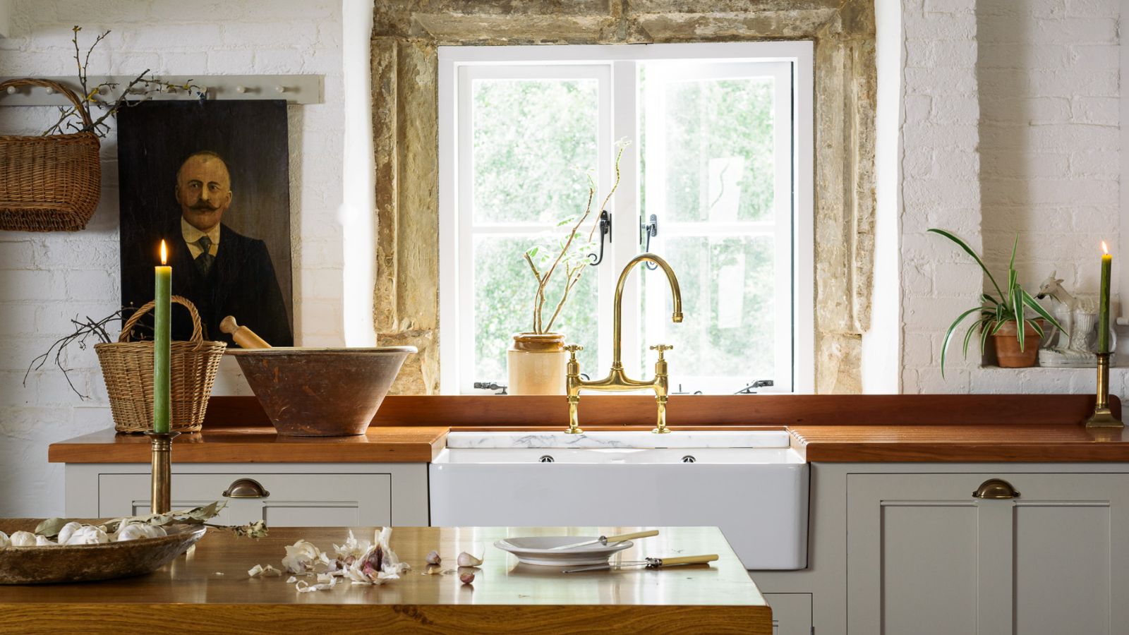 country kitchen with neutral cabinets, wooden countertop, white painted exposure stone walls with a stone window reveal and vintage decor pieces including an antique oil painting of a man
