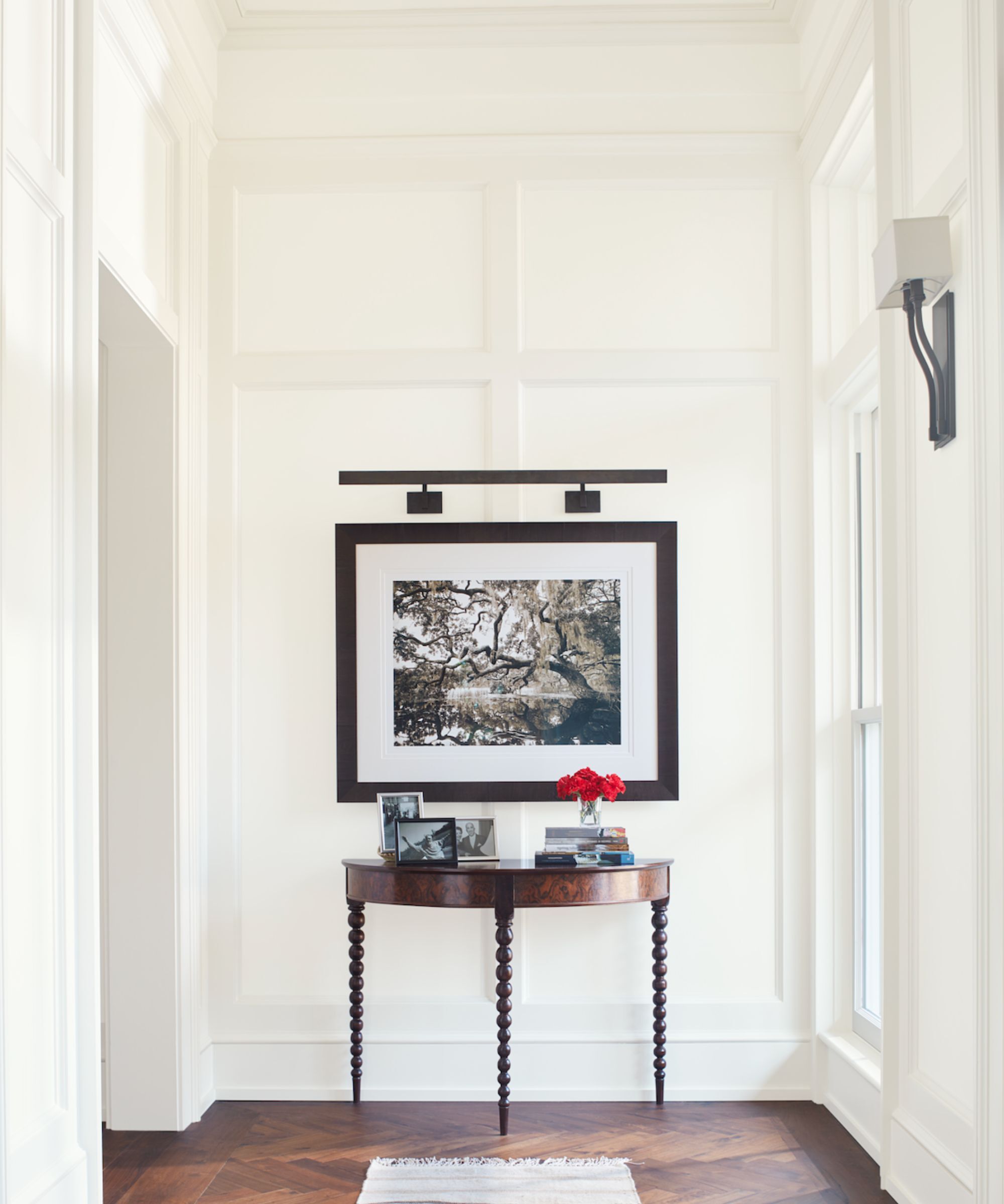 A large hallway with panelled walls painted in Benjamin Moore, Simply White. There is an antique table at the end of the hallway with a large framed photograph and picture light.