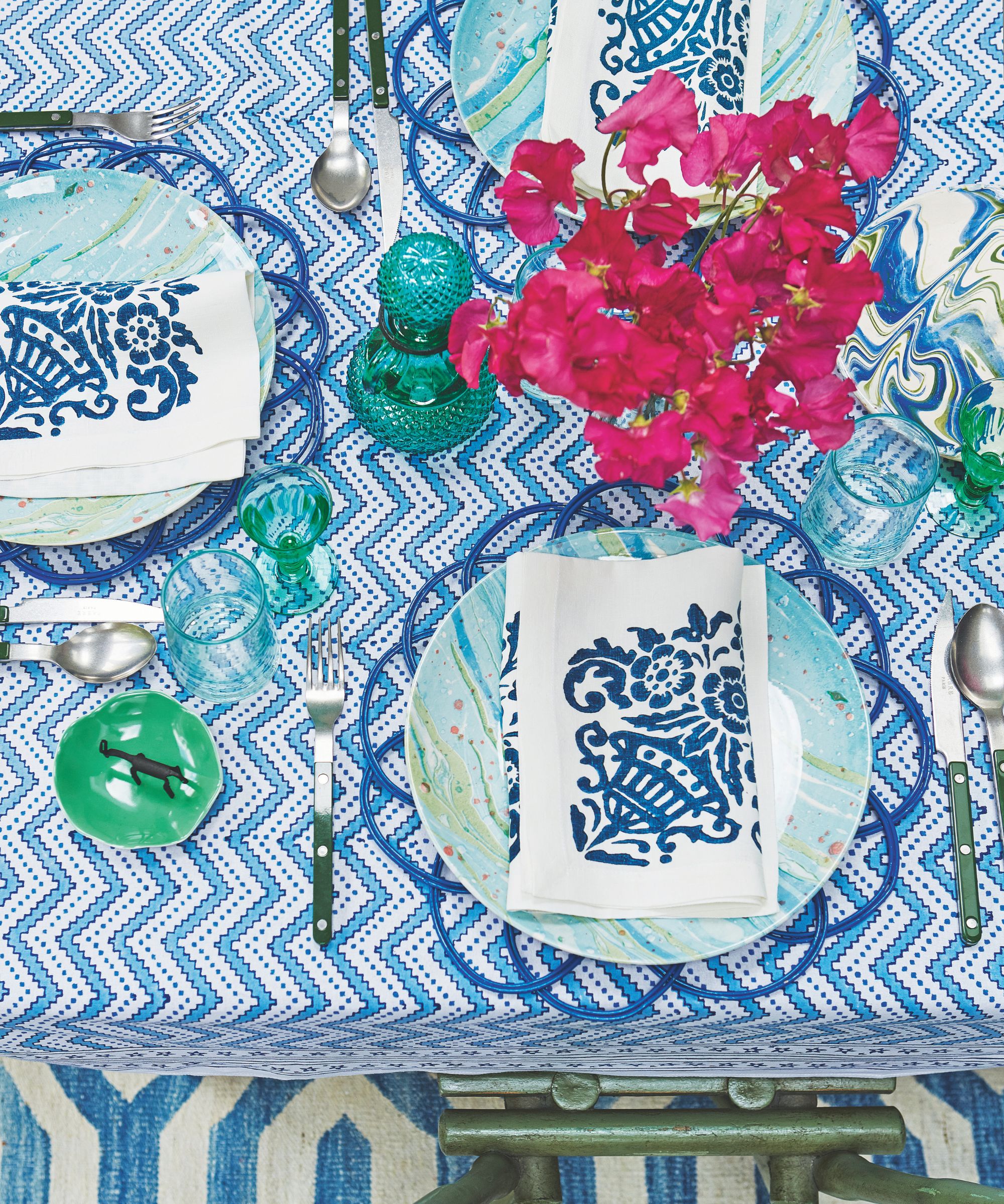 overhead shot of a table setting with blue and white zig zag tablecloth, plates and napkins with blue and white mixed prints and a vase of strong pink sweet peas
