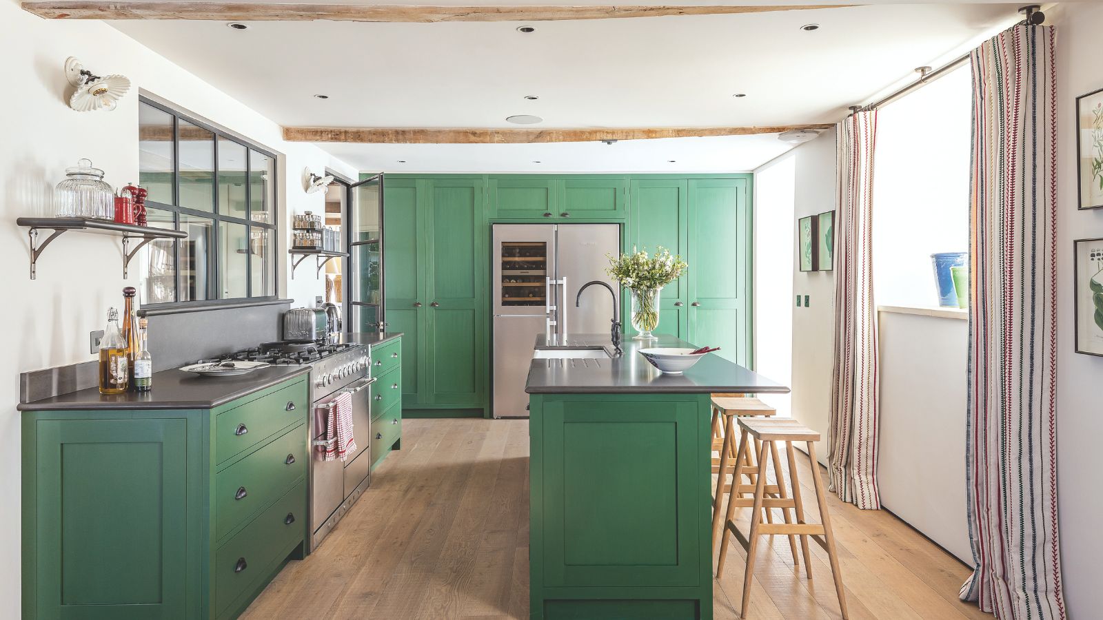Kitchen and dining area with green cabinets, beamed ceilings, paneled window above gas range, kitchen island with faucet and flower vase next to high stools. On hardwood floors in well-lit room.