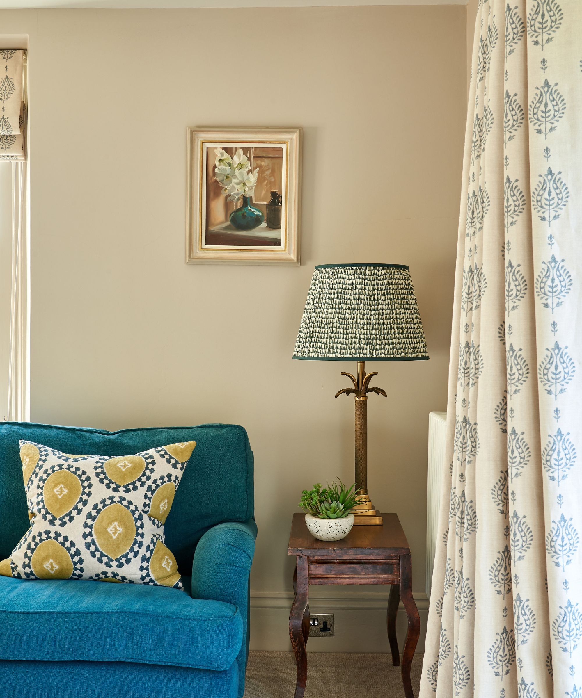 The corner of a sitting room with a bright blue sofa with a small side table on which sits a brass table lamp, the walls are painted in Slaked Lime by Little Greene