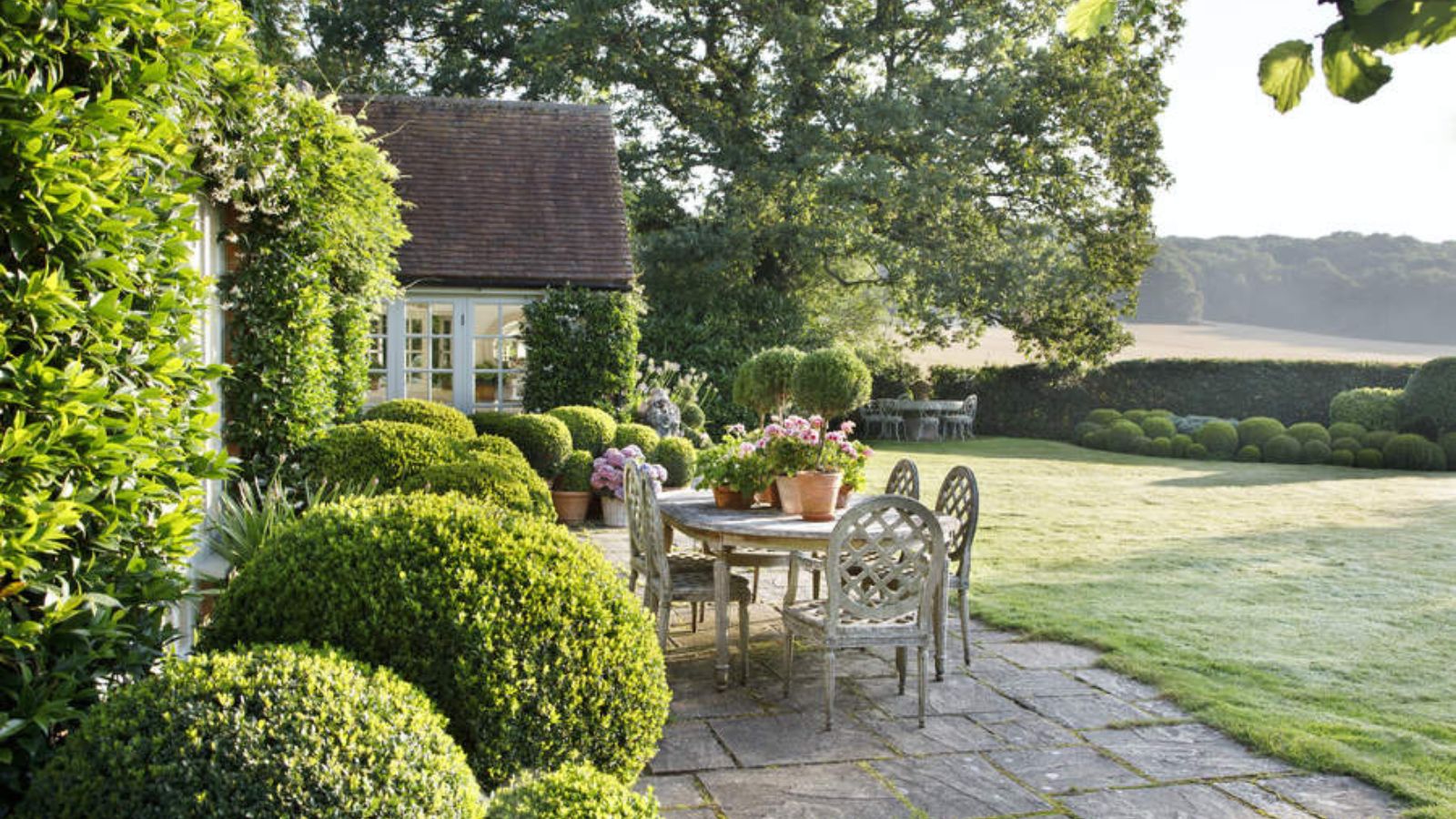 A patio furniture set in front of a cottage, surrounded by lush greenery. 