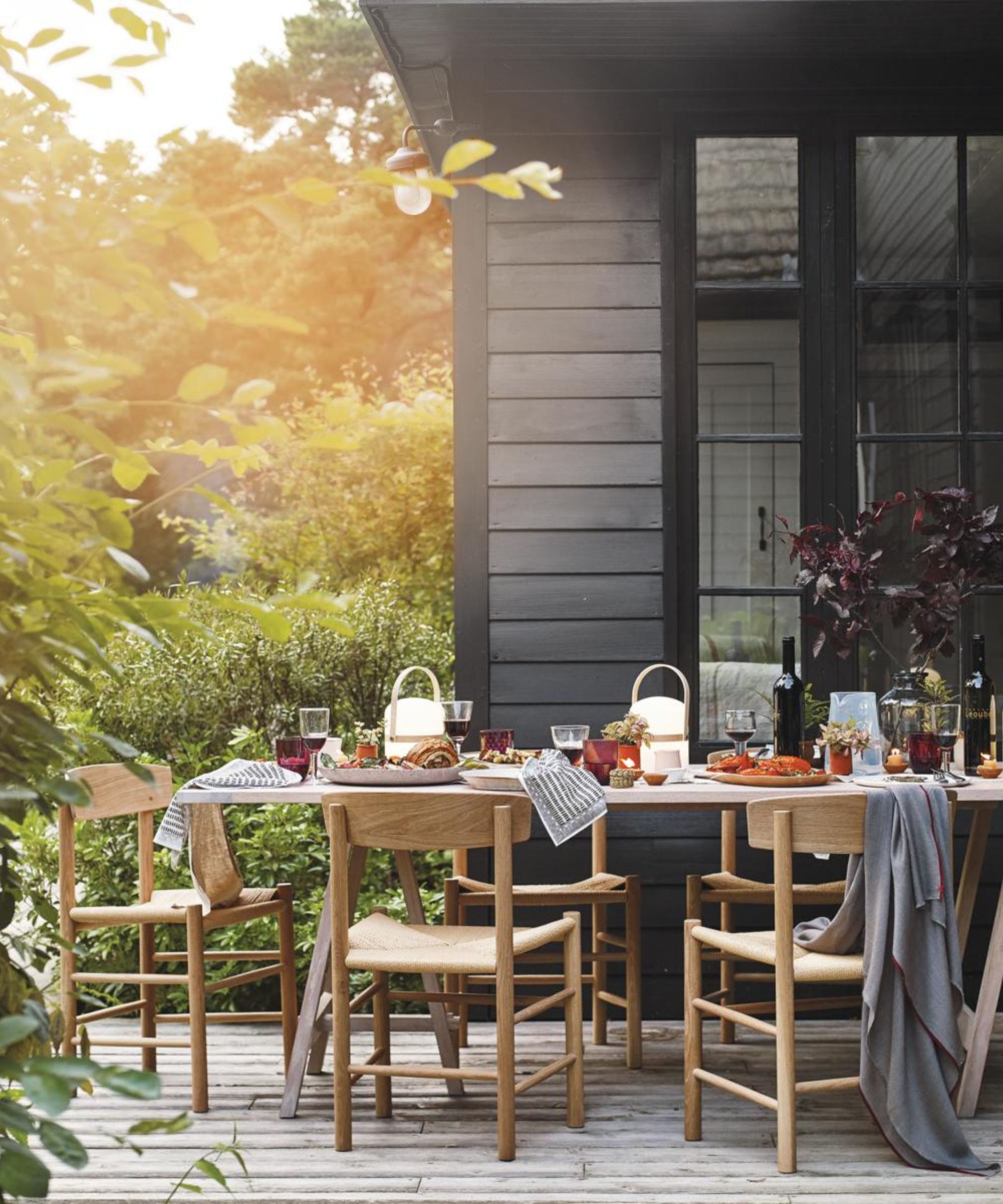 A wooden table and chairs on a deck in the setting sun, set with food and drinks, with greenery in the foreground and a black wood-clad building behind