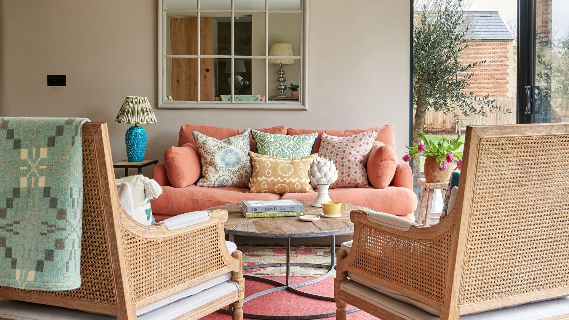 A sitting room with walls in Portland Stone by Little Greene, there is an orange sofa with colorful scatter cushions, and two rattan armchairs with a blue throw.