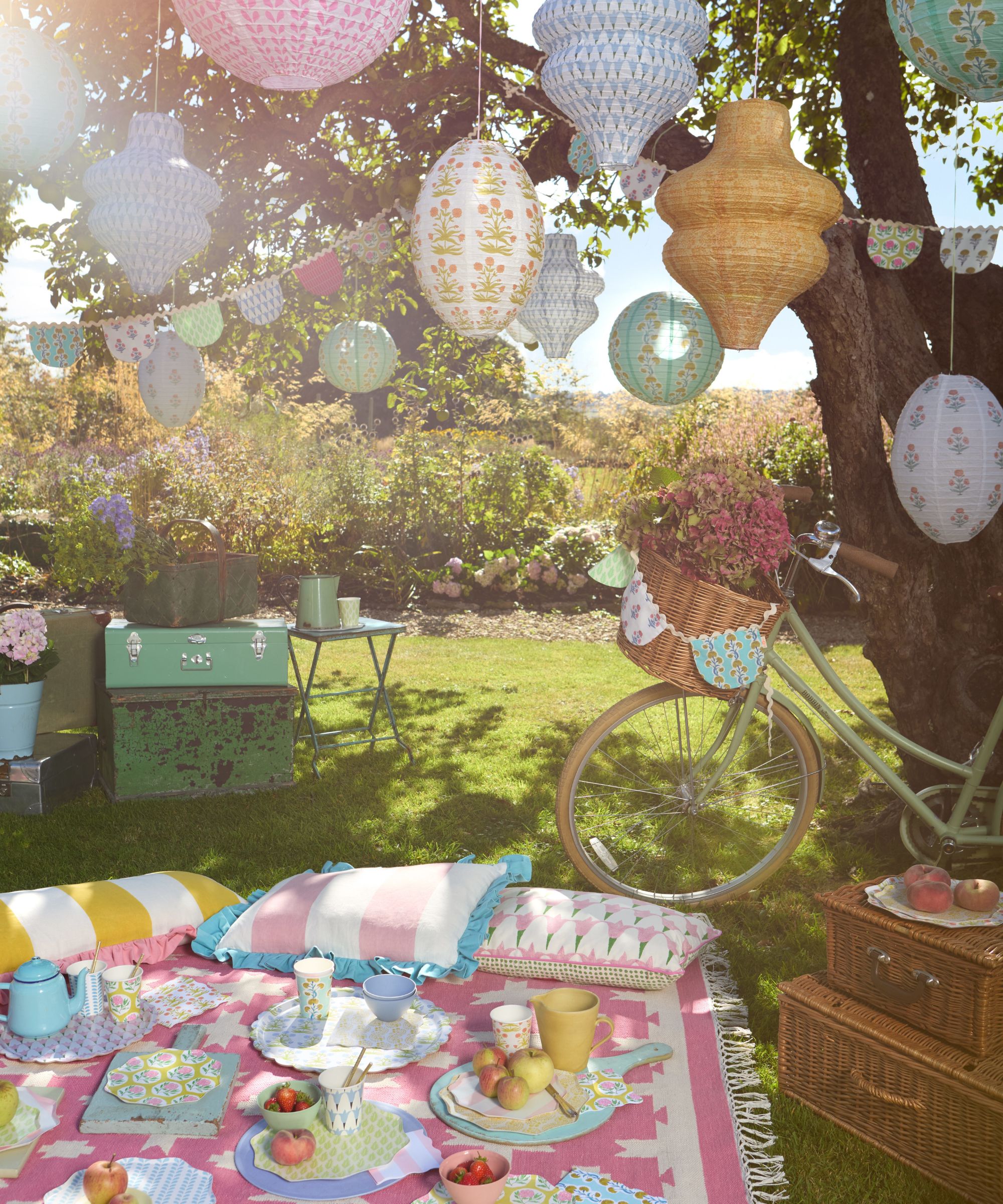 Pretty garden set up with pastel picnic blanket and cushions and paper lanterns hanging in trees, with a bike and hampers on the grass
