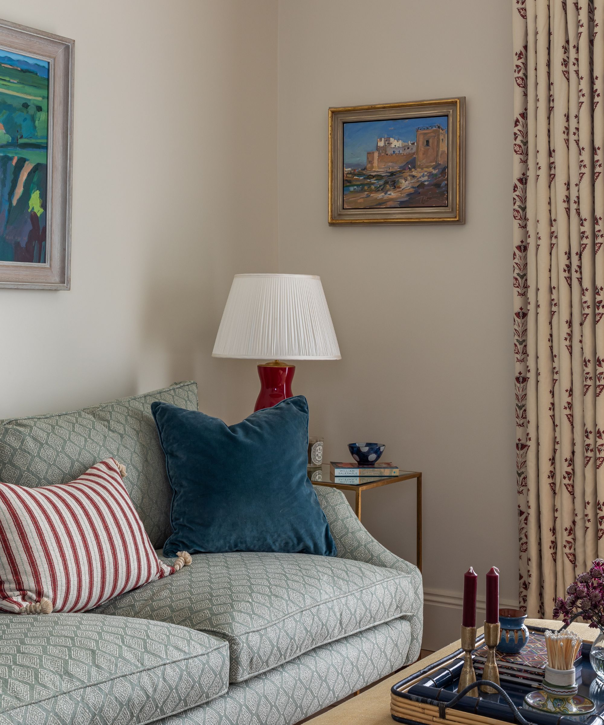 The corner of a living room with creamy beige walls painted in Slipper Satin by Farrow & Ball. There is framed artwork on the walls and the living room features a pale blue sofa with scatter cushions, a red lamp with the white pleated lampshade and a coffee table with a series of decorative items on a tray