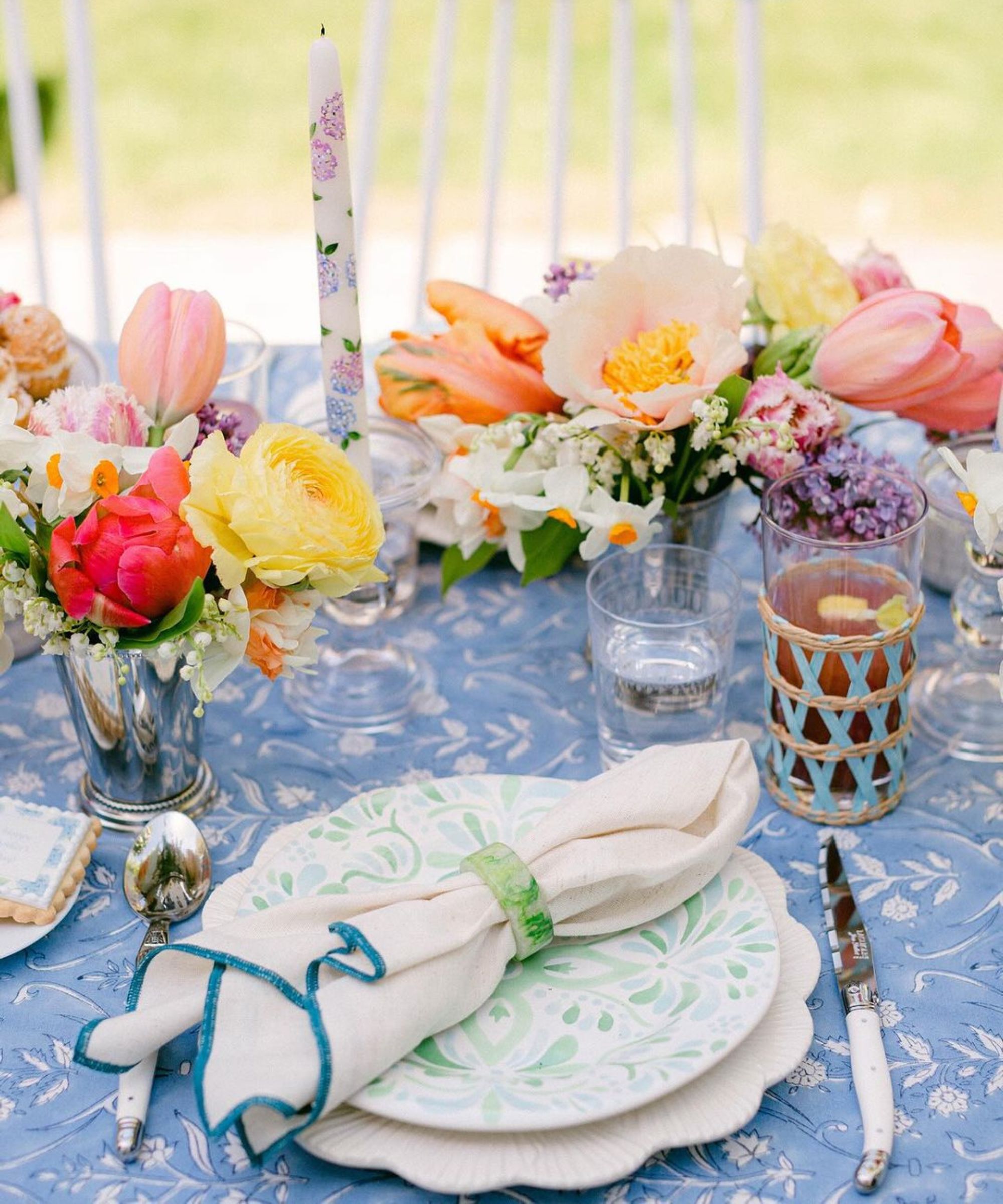 table place setting outdoors with blue tablecloth, scalloped plate, multicoloured flowers in small vases and candles