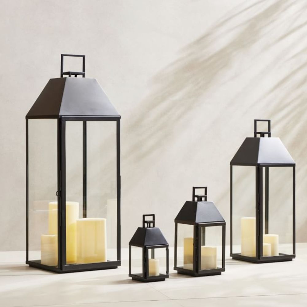 Four different sized black and glass hurricane lanterns with pillar candles on neutral background