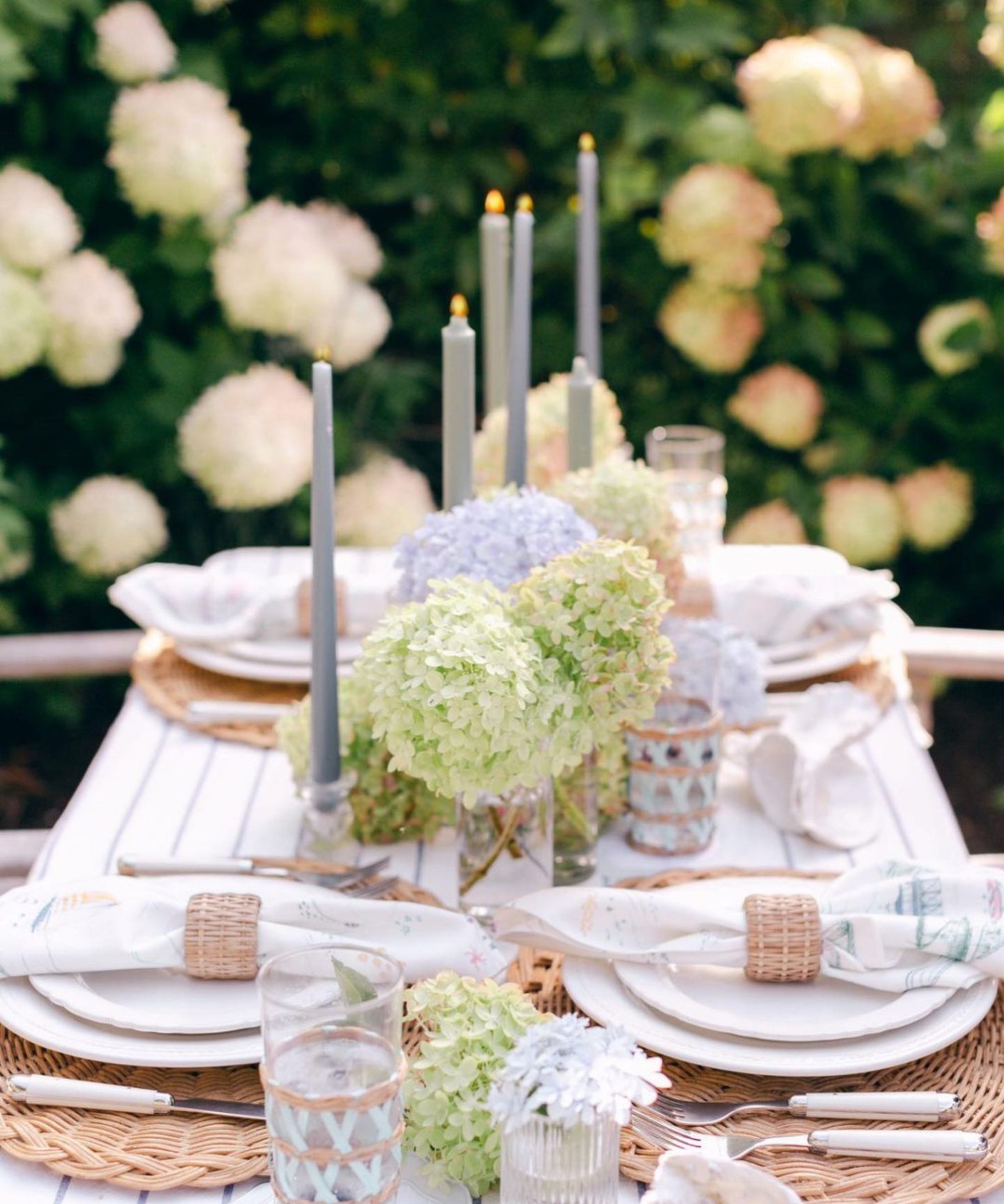 White wooden table set with white plates and rattan placemats and napkin rings, with a white hydrangea centerpiece and tall candles