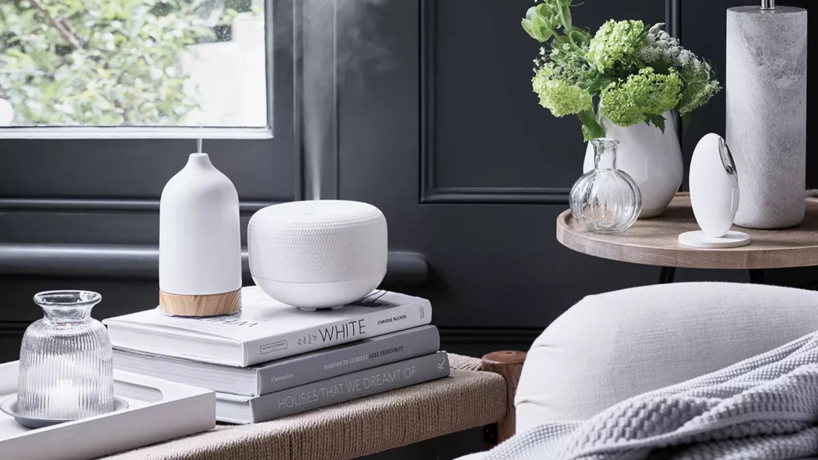 The White Company Essential Oil Diffuser on a stack of books beside a white armchair against gray walls. 