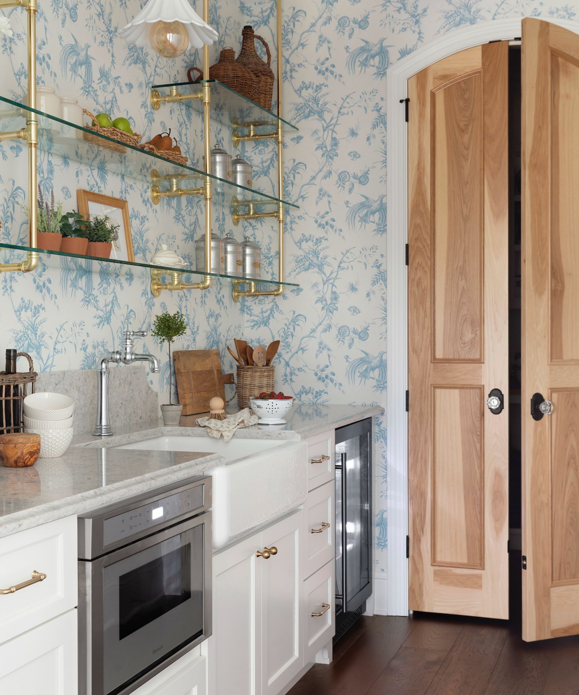 A small kitchen with white cabinets painted in Simply White by Benjamin Moore, walls in a blue and white wallpaper and glass shelving.