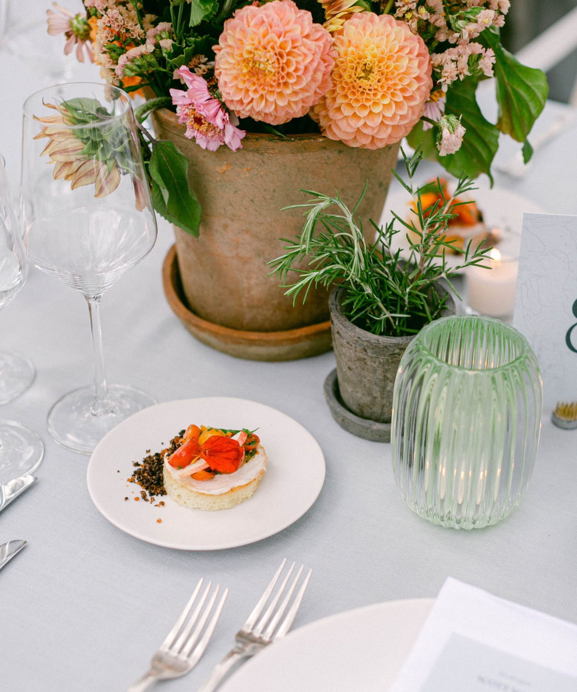 Terracotta pot of orange and pink flowers on a table with white tablecloth and a desert on a plate with wine glasses and candle holders