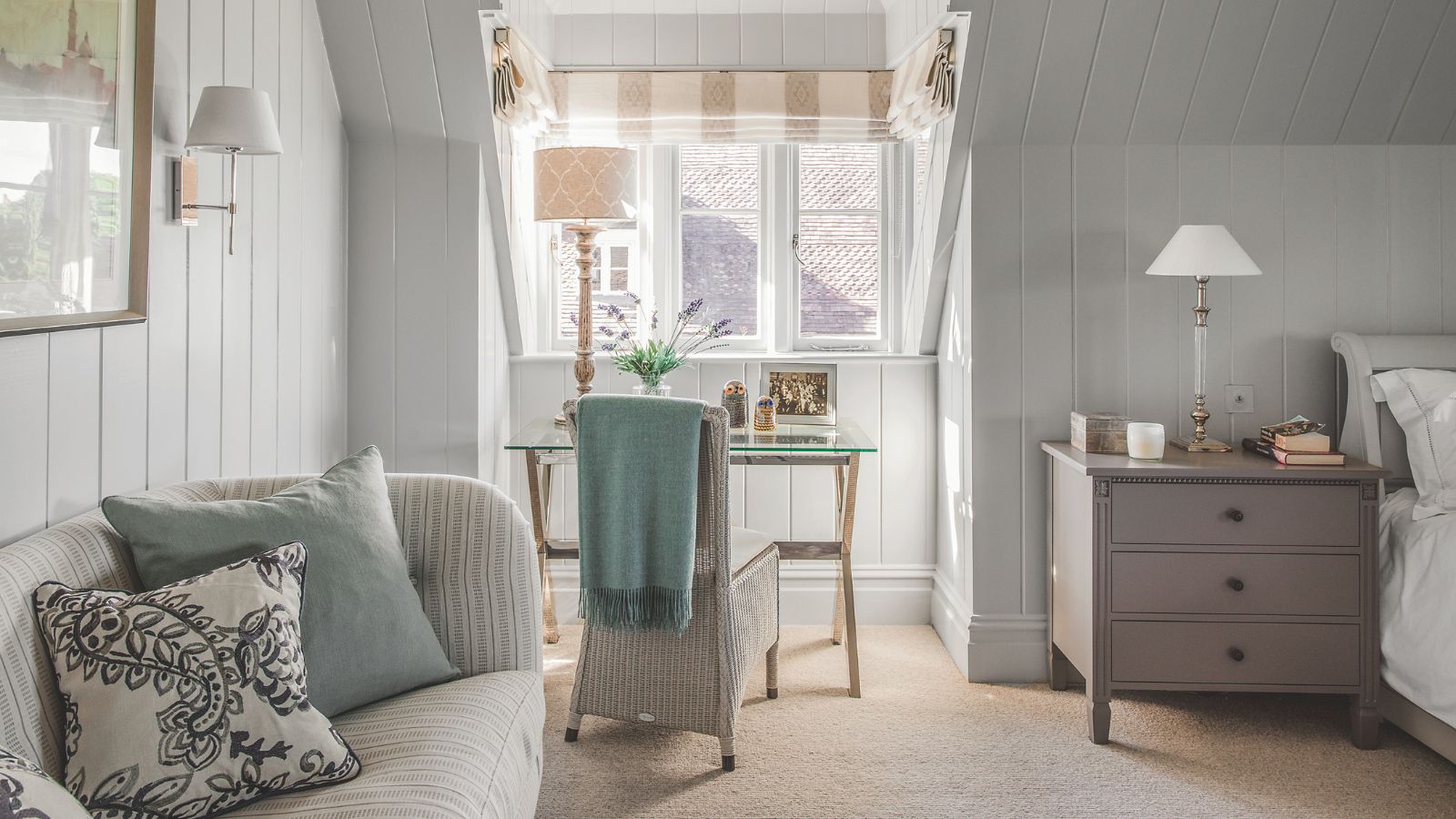 Cozy bedroom with wood panelled walls in light grey, a glass dressing table near a window and a sofa with scatter cushions in pale green, natural light through window.