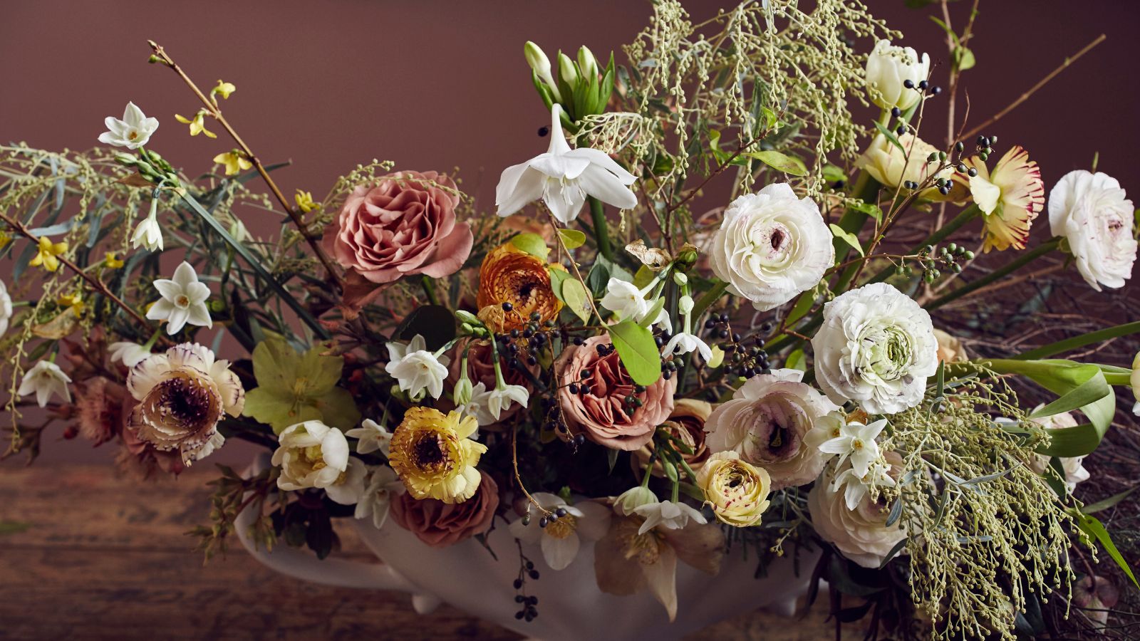 bouquet of flowers in rust, pink and white colours in a large white handled vase on a wooden surface and dusky pink painted background