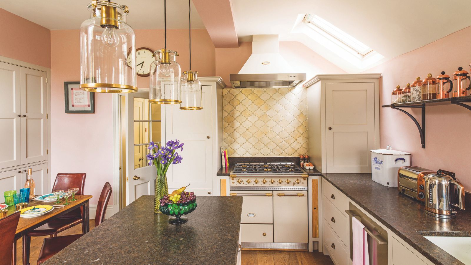 Kitsch pink kitchen with yellow shel-shaped backsplash tiles behind cream and gold range cooker. It's a kitchen diner with breakfast bar and gold accented pendant lights
