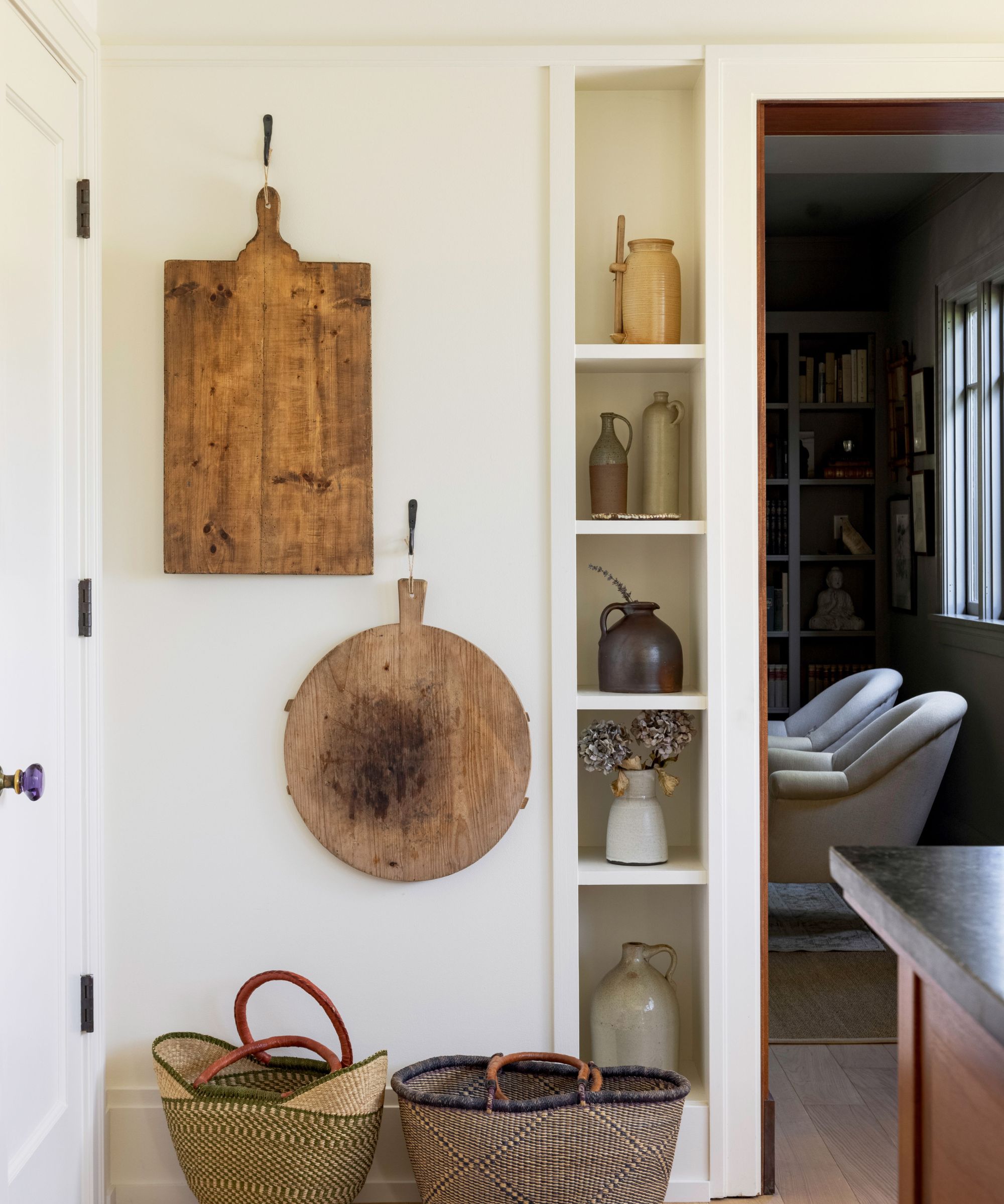 A corner of a kitchen painted in Swiss Coffee by Benjamin Moore. The wall has hanging items, including chopping boards and baskets, as well as narrow display shelves and wooden floors.