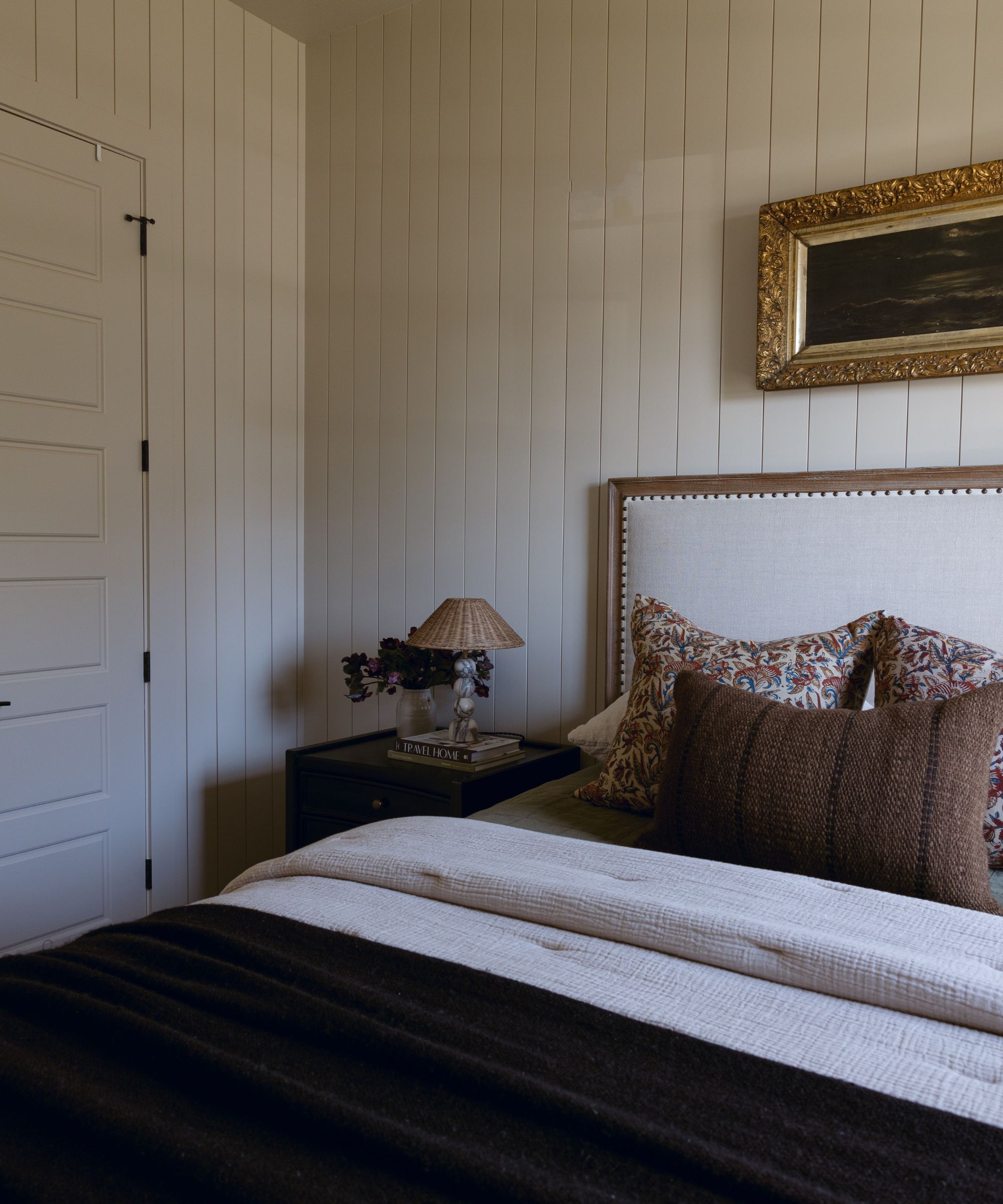 A bedroom with panelled walls, painted in Accessible Beige by Sherwin-Williams. The bed has a beige upholstered bedframe and cream bedding with brown and dark beige colored pillows and throw.