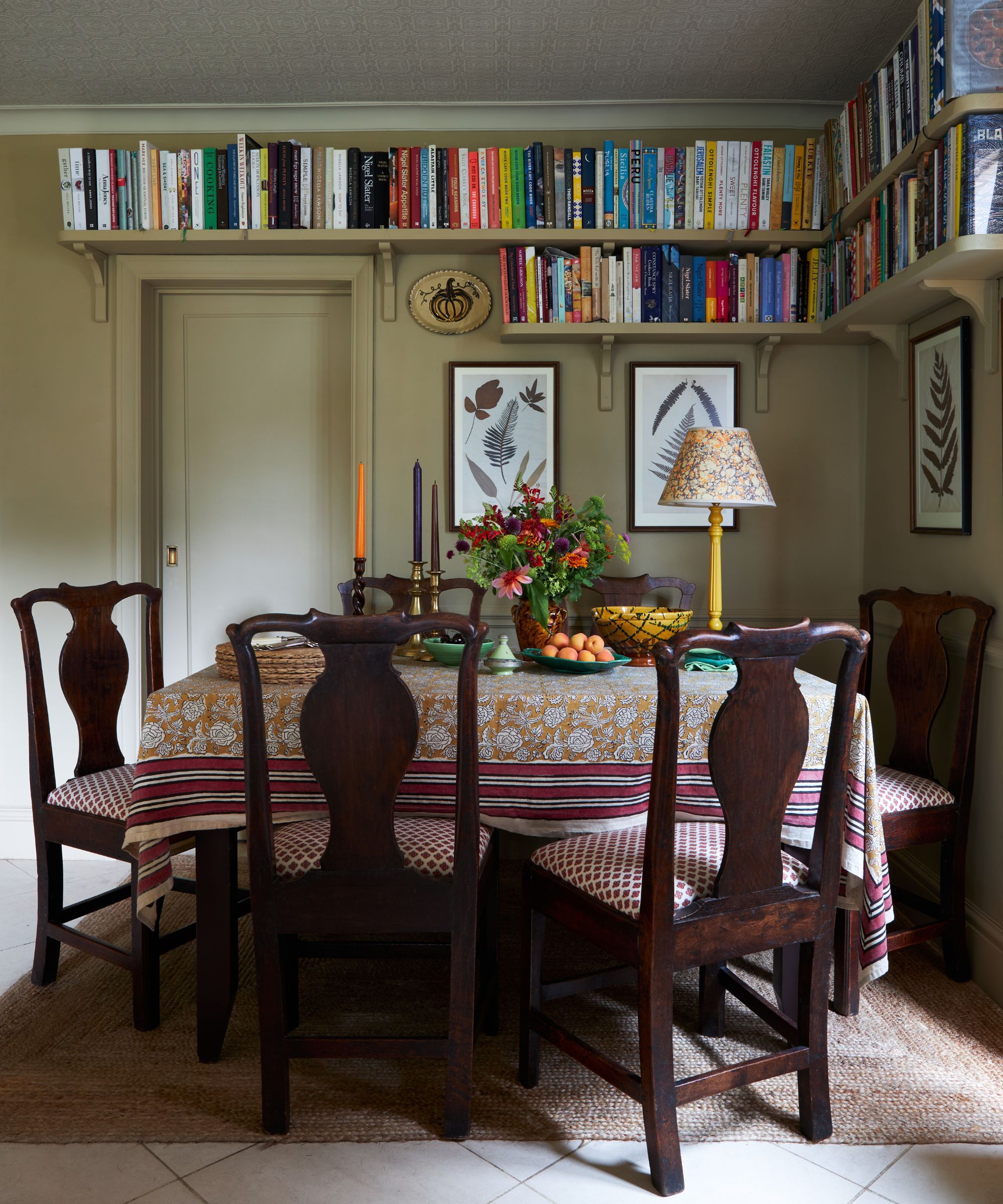 A cottage style dining room painted in Biscuit by Farrow & Ball, there is a traditional dining table with an orange and red table cloth and mahogany dining chairs. There is shelving high on the walls filled with books.