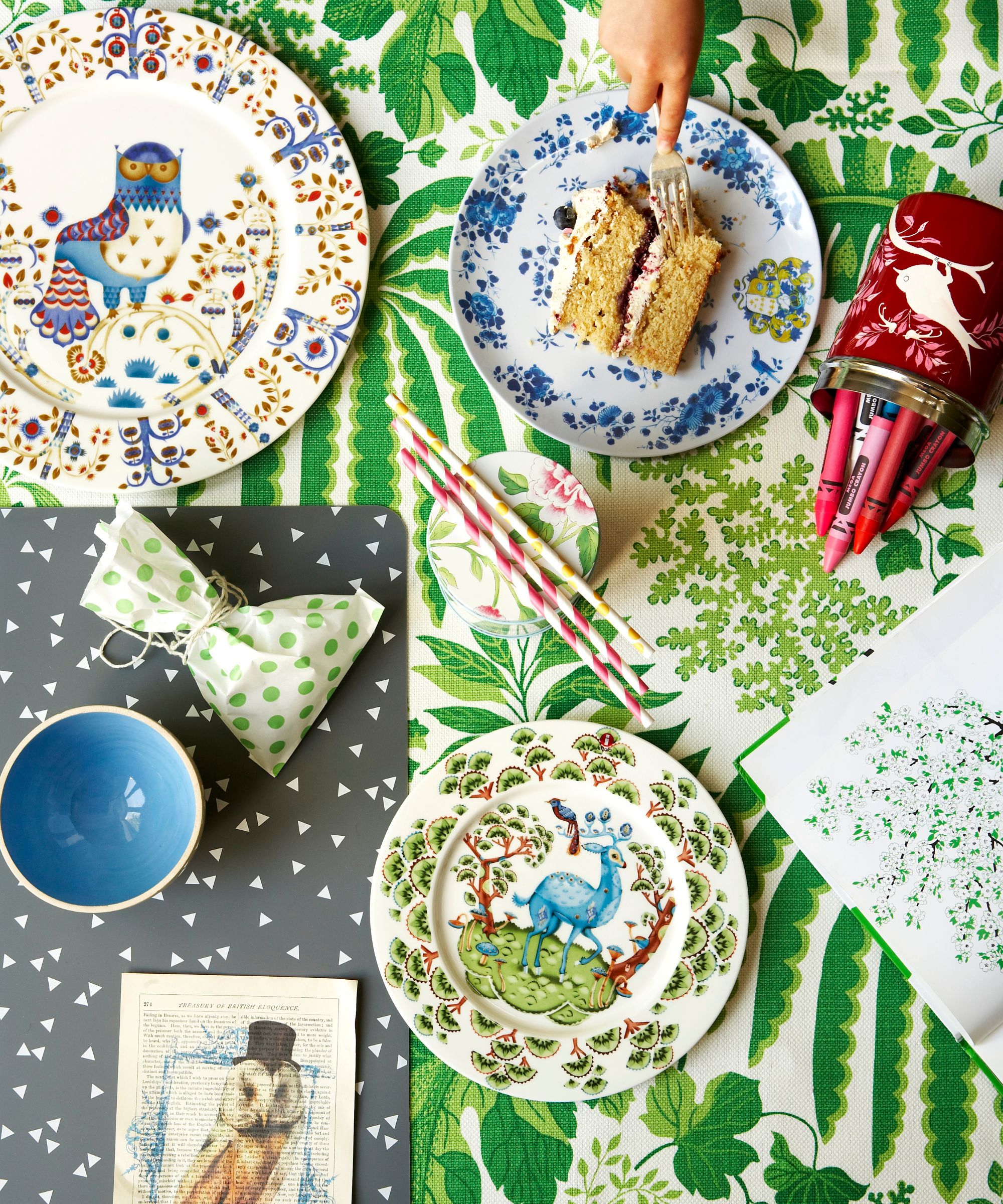 overhead shot of a table with a leafy green tablecloth, decorative plates, a child's cutting some cake and some crayons, striped straws and polka dot gift bag